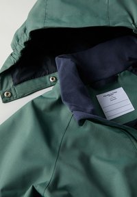 Groene jas met een marinekleurige kraag, voorzien van een ritsluiting en drukknoopsluiting. Binnenlabel zichtbaar, met merk- en maatinformatie.