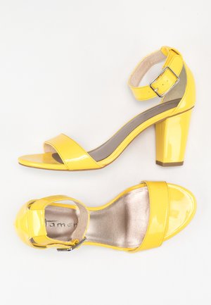 Gule patentlæder hæle med blokhæle, ankelstrop og åbent tå. Har en blank overflade og polstret indersål for komfort.