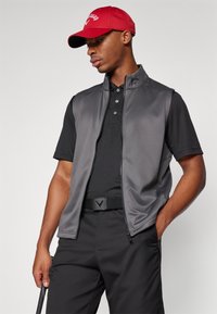 Un gilet sans manches gris sur un polo noir, accompagné de pantalons noirs. Une casquette rouge arbore un logo blanc. L'ensemble est élégant et moderne.