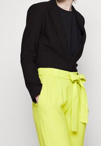 Czarna marynarka z głębokim dekoltem w kształcie litery V, zestawiona z jasnymi, żółtymi spodniami z wysokim stanem, wyposażonymi w wiązany pasek i boczne kieszenie. Tkanina o fakturze.