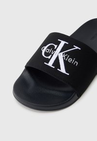 Zwarte slide-sandalen met een gestructureerde strap met daarop het witte geborduurde "CK"-logo. Zacht rubberen zool; casual, open teen ontwerp.