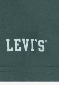 Tmavě zelená bavlněná tkanina s bílým logem "LEVI'S®". Hladká textura se zpevným stehem na lemu. Jednoduchý, klasický design.