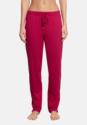 Kobieta w jasnoróżowych spodniach dresowych z troczkiem, stojąca boso z rękami swobodnie opuszczonymi wzdłuż ciała na jednolitym białym tle.