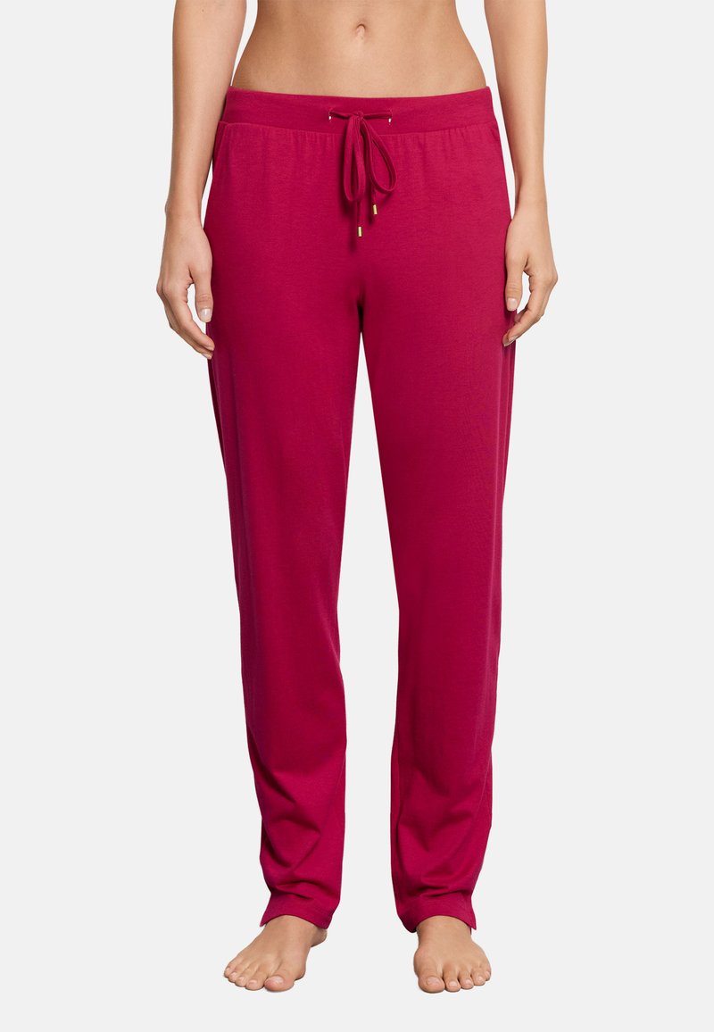 Femme portant un pantalon de détente rose vif à cordon, debout pieds nus avec les mains détendues le long du corps sur un fond blanc uni.