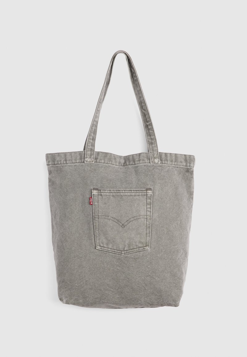 Sac fourre-tout en denim gris avec double poignée et une poche carrée cousue ornée d'une petite étiquette rouge sur un fond uni.