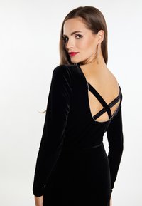 Schwarzes Samtkleid mit langen Ärmeln, tiefem V-Ausschnitt im Rücken, gekreuzten Trägern und dekorativen silbernen Akzenten am Halsauschnitt.
