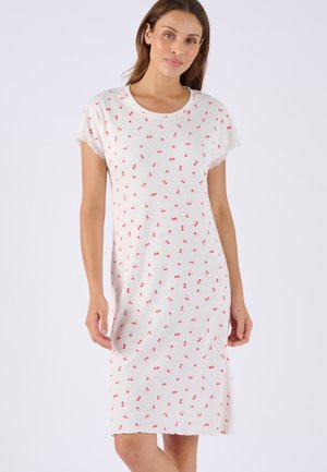 Frau trägt ein weißes knielanges Kleid mit kleinen roten Kirschmotiven und Spitzenbesatz an den kurzen Ärmeln, steht vor einem schlichten Hintergrund.
