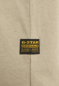 Tessuto beige con etichetta rettangolare nera con testo giallo: "G-STAR ORIGINALS RAW | DENIM" centrato lungo la cucitura verticale.