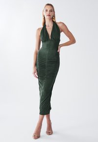 Zelena obleka s halter ovratnikom, globokim V izrezom, oprijetim krojem in nabranim vzorcem spredaj, dolžine do kolen ter mehkim materialom.