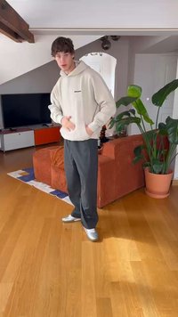 Lichtgrijze, gestructureerde hoodie met een voorzak, gecombineerd met donkere broeken en zilveren sneakers. Op de achtergrond staat een potplant en een oranje bank.