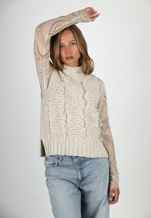 Frau mit schulterlangem Haar, die eine weiße, strukturierte Strickweste über einem gemusterten, transparenten Langarmshirt und hellblauen Jeans trägt, mit einem erhobenen Arm.