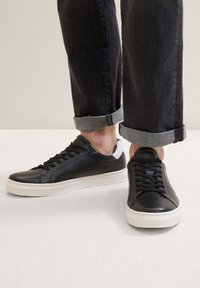 Schwarze Ledersneaker mit weißen Gummisohlen, runden Zehen, flachen Schnürsenkeln und einem strukturierten Design; getragen mit umgeschlagenen dunklen Jeans.
