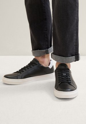 Baskets en cuir noir avec semelles en caoutchouc blanches, dotées de bouts ronds, de lacets plats et d'un motif texturé ; portées avec un jean foncé retroussé.