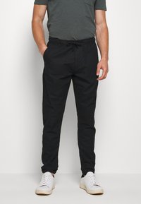 Schwarze, tapered Hosen aus leichtem Stoff mit elastischem Bund und Kordelzug. Kombiniert mit weißen Turnschuhen und einem dunkelgrauen Shirt.