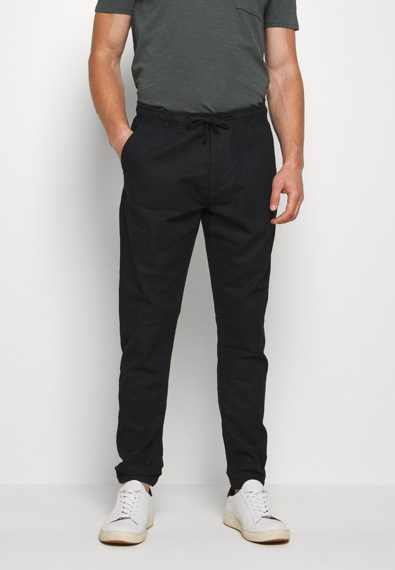 Schwarze, tapered Hosen aus leichtem Stoff mit elastischem Bund und Kordelzug. Kombiniert mit weißen Turnschuhen und einem dunkelgrauen Shirt.