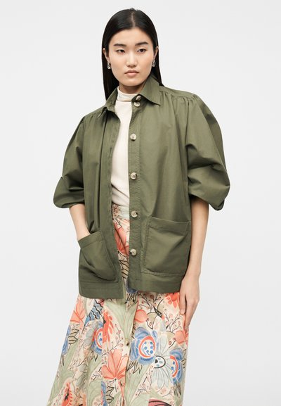 Γυναίκα με μακριά σκούρα μαλλιά φοράει πράσινο μπουφάν με κουμπιά και μεγάλες τσέπες, μπεζ τοπ και πολύχρωμη φλοράλ φούστα, στέκεται με τα χέρια μερικώς στις τσέπες.