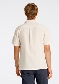 Camisa de manga corta, color beige claro, con un corte relajado, que presenta un pliegue en la parte trasera y un diseño de cuello simple, confeccionada en una tela texturizada.
