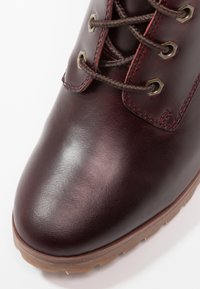 Botte en cuir marron foncé avec un bout rond, des lacets marron passant à travers des œillets en laiton, et une semelle en caoutchouc marron clair.