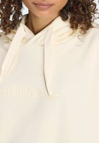 Krémová mikina s kapucňou a textúrovanými šnúrkami, na prednej strane s vyvýšeným, tónovým logom adidas a emblémom trojlístka. Mäkký materiál.