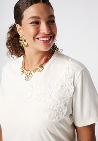 T-shirt blanc à manches courtes avec des appliqués floraux texturés, associé à un collier en chaîne doré avec divers charms et de grandes boucles d'oreilles dorées.