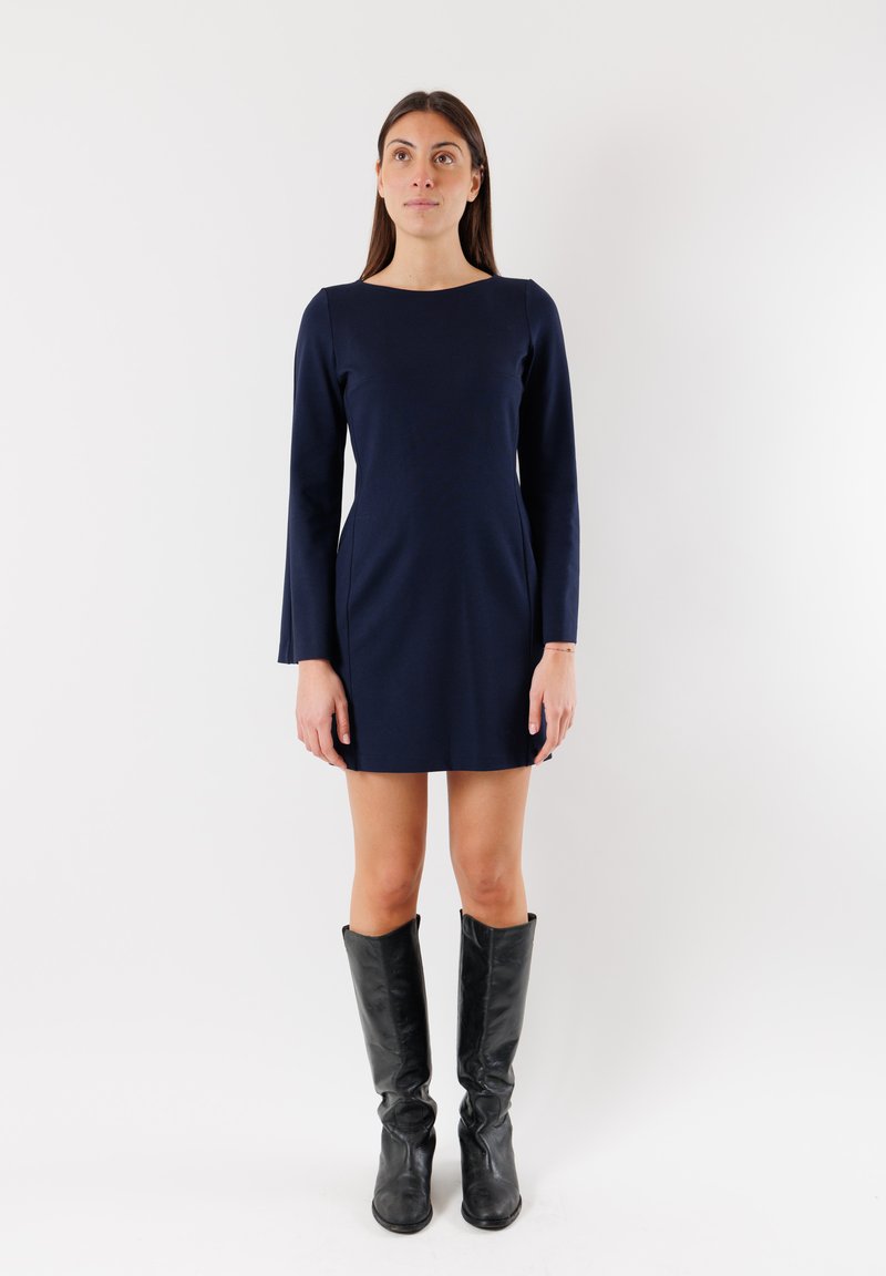 Abito lungo blu navy con maniche lunghe, scollatura a barca, silhouette aderente e texture liscia, abbinato a stivali in pelle neri al ginocchio.