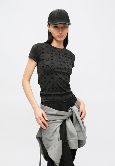 T-shirt preta de manga curta com um padrão circular subtil, combinada com um casaco cinzento amarrado à cintura e um boné preto.