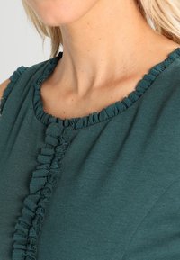 Turkusowa bluzka z okrągłym dekoltem, ozdobiona falbaniastymi akcentami wzdłuż kołnierza i przeszycia z przodu, wykonana z fakturowanej tkaniny.