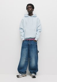 Hoodie oversize azzurro con tasca frontale, abbinato a jeans in denim blu larghi con dettagli in patch e etichetta. Completato con scarpe sportive.