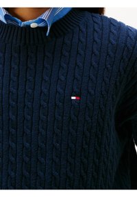 Pull en maille côtelée bleu marine avec un col côtelé et un petit logo rouge, blanc et bleu en accent. Le tissu présente des variations visibles de texture et de motif.