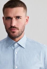 Mann mit kurzen braunen Haaren und Bart, der ein hellblau kariertes Hemd mit Knöpfen vor einem schlichten grauen Hintergrund trägt.