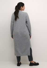Cardigan lungo grigio a coste con alte fessure ai lati, abbinato a pantaloni scuri e scarpe slip-on nere.