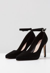 Svarta mocka högklackade skor med spetsiga tår och ankelremmar med spänne. Klackhöjden är cirka 10 cm. Slät innersula.