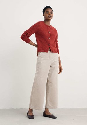 Vrouw staat met hand op de heup, gekleed in een roestrode knoopcardigan, beige wijde broek en donkere platte schoenen tegen een witte muur.