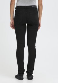 Personne portant un jean noir ajusté et des chaussures noires, debout de dos contre un fond blanc uni.