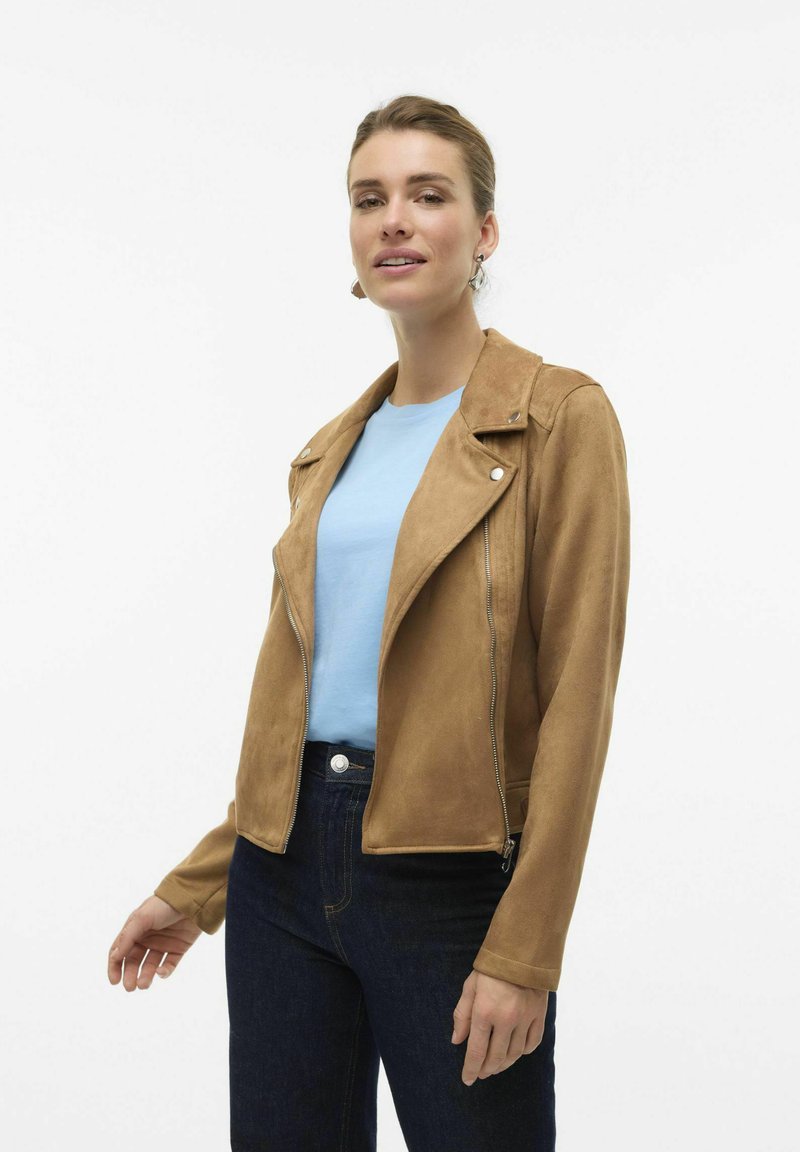 Veste de moto en daim marron au design court, avec quincaillerie argentée et devant zippé, associée à un haut bleu clair et un jean foncé.