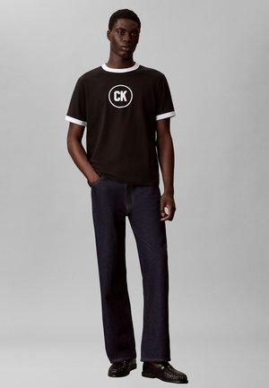 Tânăr purtând un tricou negru cu logo-ul alb „CK” și detalii albe, blugi închiși la culoare și pantofi negri, stând cu o mână în buzunar.