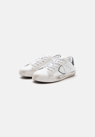 Baskets basses blanches avec des empiècements en daim gris, des languettes noires au talon, des lacets imprimés, des semelles usées et sales, et un logo en forme de bouclier sur le côté.