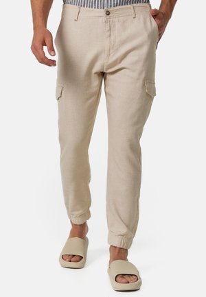 Homme portant un pantalon jogger cargo beige avec des poignets élastiques et des sandales slides beiges assorties, debout devant un fond blanc.