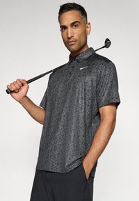 Pánská polokošile s krátkým rukávem v tmavě šedé barvě s květinovým vzorem, s límečkem na knoflíky a logem Nike. Model drží golfovou hůl.