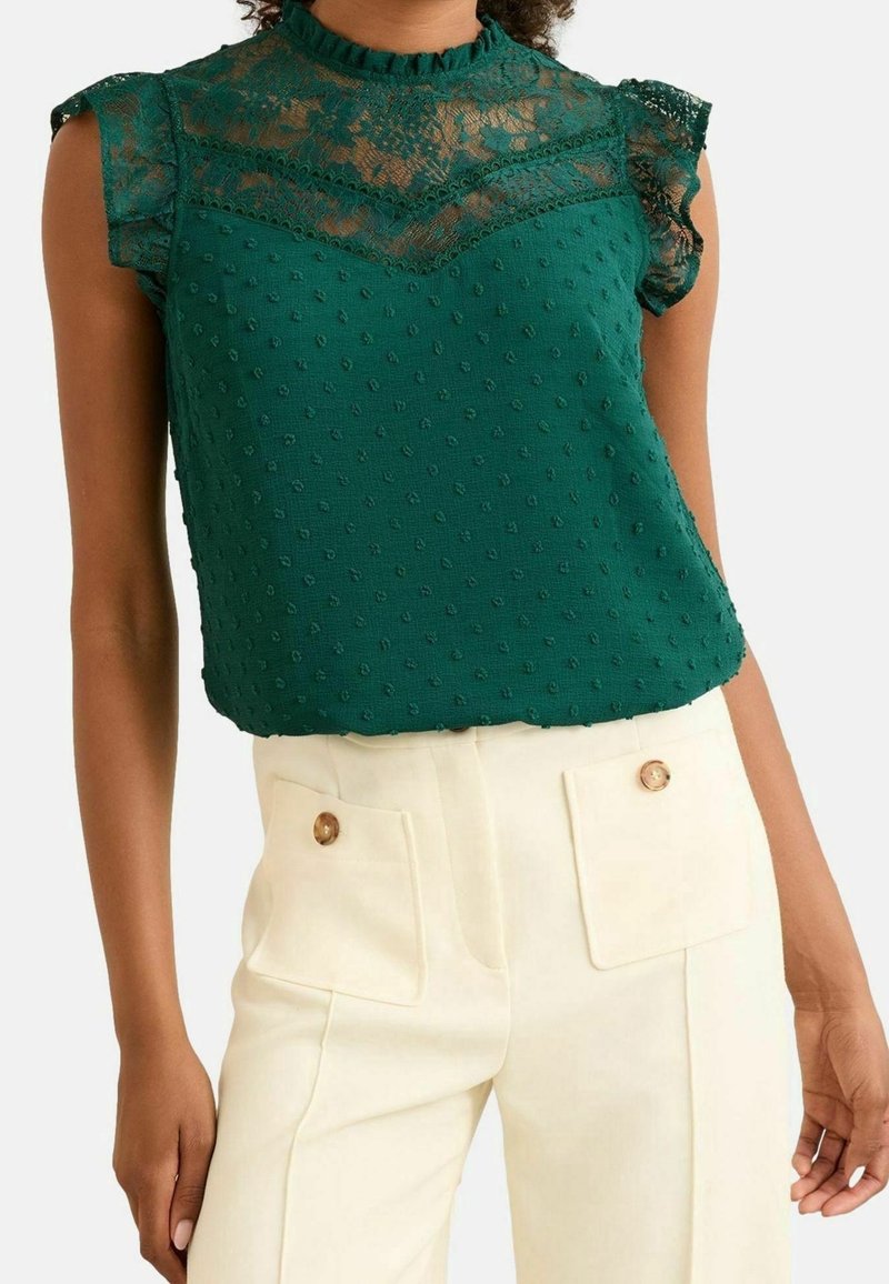 Blouse verte sans manches en dentelle avec des pois texturés, associée à un pantalon crème taille haute avec poches avant boutonnées.