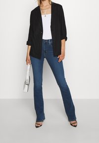Černý blazer s ohrnutými rukávy přes bílý top na ramínka, doplněný úzkými modrými zvonovými džínami. Doplněno texturovanou stříbrnou kabelkou.
