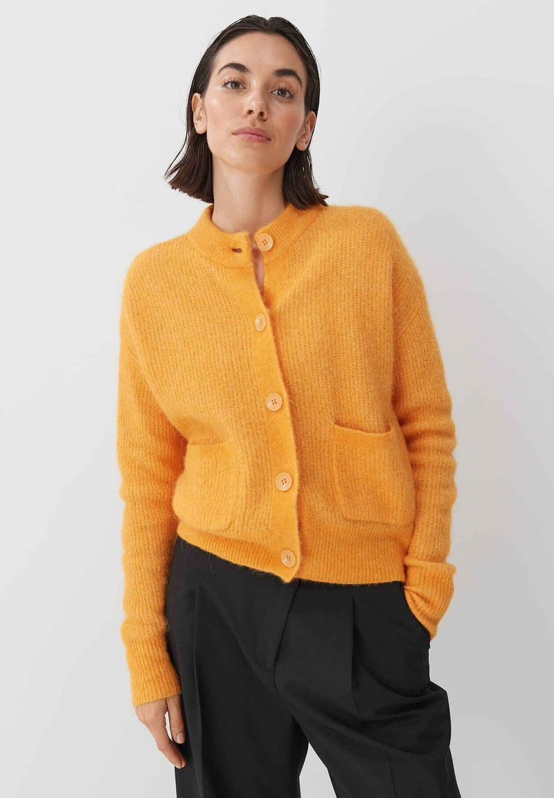 Orange cardigan with a round neckline, ribbed texture, five button closure, and two front pockets. Long sleeves complement the relaxed fit.