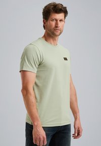 Svetlozelené tričko s krátkym rukávom a okrúhlym výstrihom, s malou čiernou nášivkou na hrudi, vyrobené z hladkej zmesi bavlny.