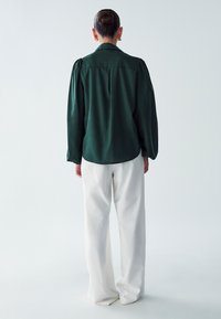 Tmavě zelená oversized košile na knoflíky se staženými rukávy, v kombinaci s bílými širokými kalhotami. Hladká textura, minimalistický design.