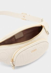 Hvid faux læder crossbody taske med et tekstureret logo-design, guld lynlåsdetaljer og en enkelt justerbar rem. Indvendig er den brun.