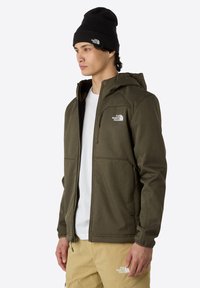 Oliwkowa kurtka typu softshell z zamkiem z przodu, regulowanym kapturem i dwiema bocznymi kieszeniami. Noszona z czarną czapką beanie i jasnobrązowymi spodniami.