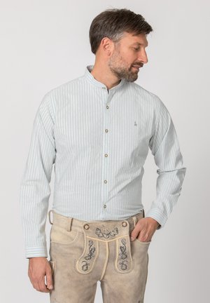 Mann trägt hell gestreiftes Hemd mit Knöpfen und traditionelle beige Lederhosen mit bestickten Blumenmustern, schaut seitlich.