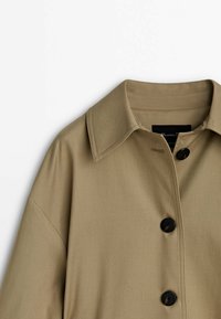 Trench-coat beige avec un large col et trois grands boutons noirs sur le devant.