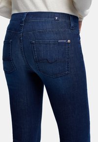 Jeans in denim blu scuro con un design aderente, dotati di tasche posteriori, cuciture sottili e un'etichetta con marchio sulla vita.
