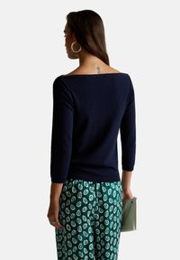 Jersey de punto marino con escote amplio, mangas de tres cuartos y dobladillo acanalado, combinado con pantalones verdes con estampado y un pequeño bolso de mano.